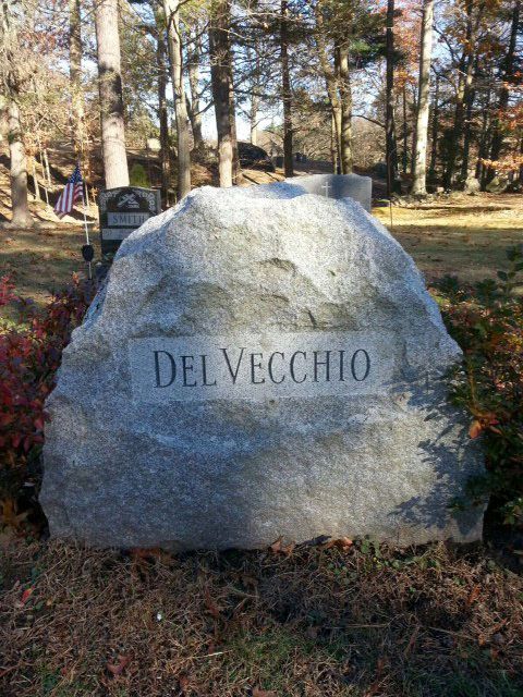 Large granite memorial stone engraved “DEL VECCHIO” in a wooded outdoor setting with autumn leaves.