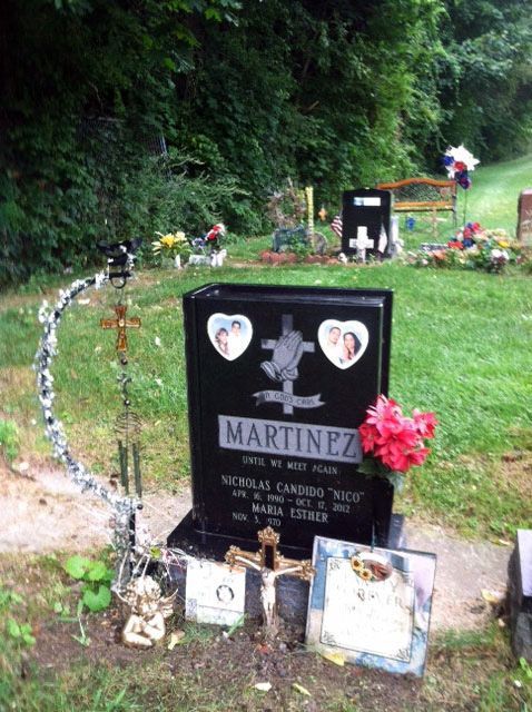 Gravestone with flowers and framed photos in a grassy cemetery beside a wooded hillside
