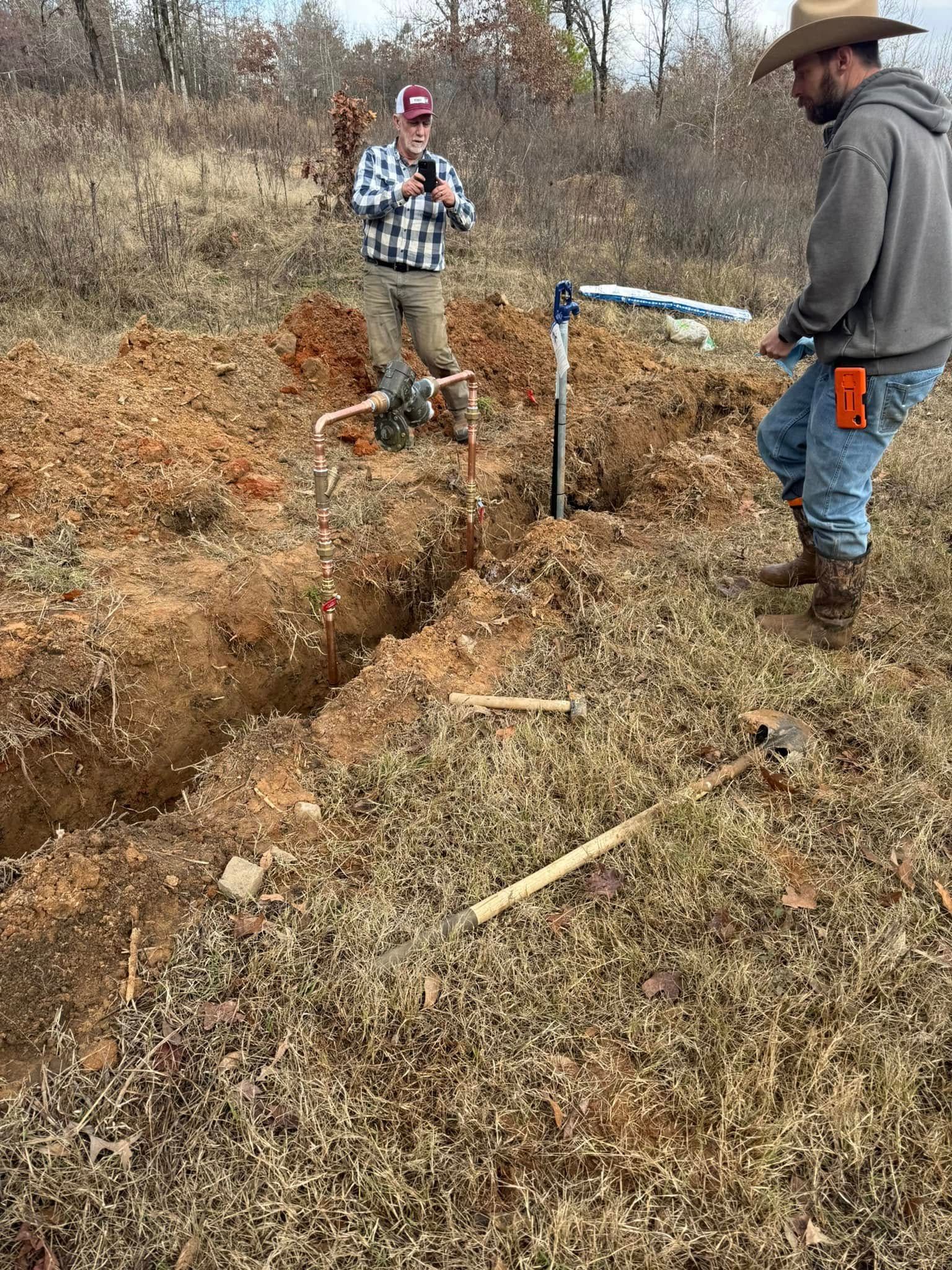 Two people standing by a freshly dug trench in a grassy field, near a blue pipe and shovel
