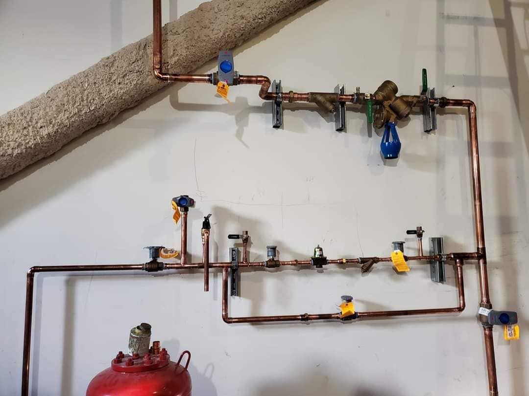 Copper plumbing pipes with blue and yellow valves on a white wall above a red tank.