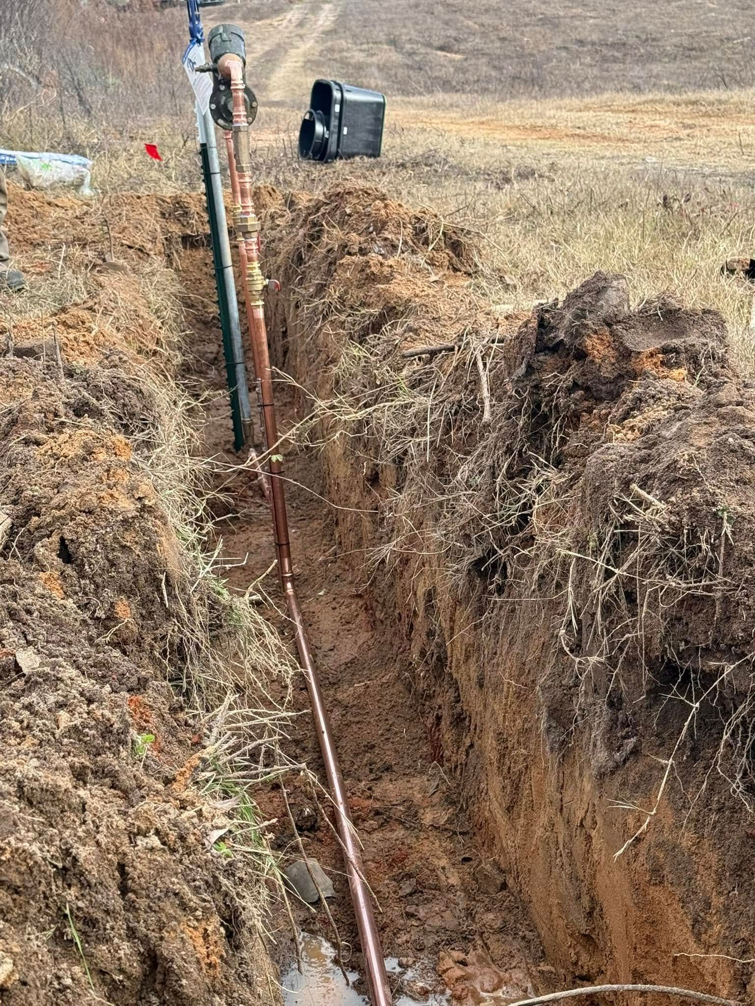 Deep trench in dry dirt with a vertical pipe and a black bucket at the edge