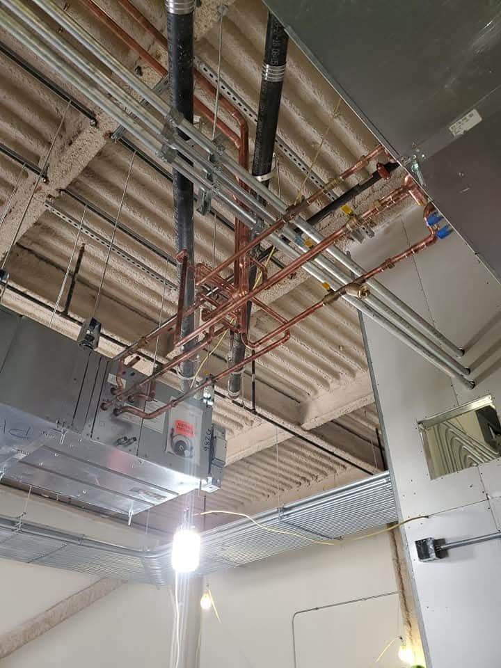 Indoor industrial ceiling with suspended pipes, cable trays, and a bright hanging light fixture.