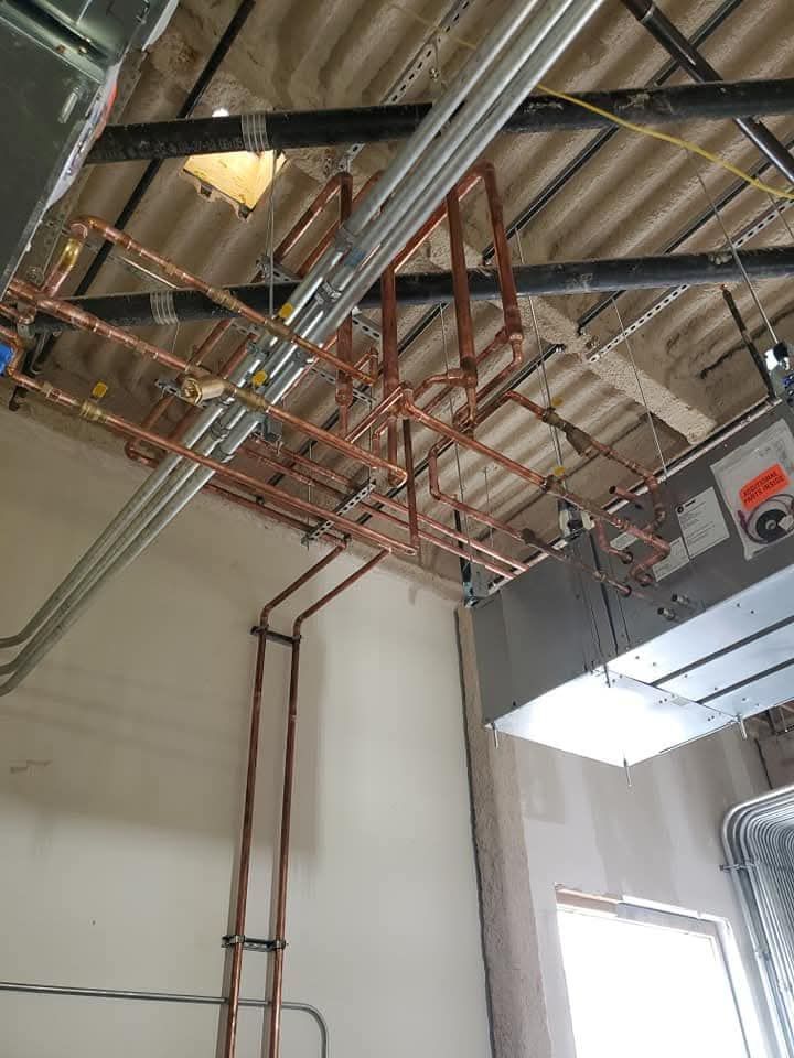 Indoor industrial ceiling with exposed pipes, ducts, and cable trays above a white wall and window