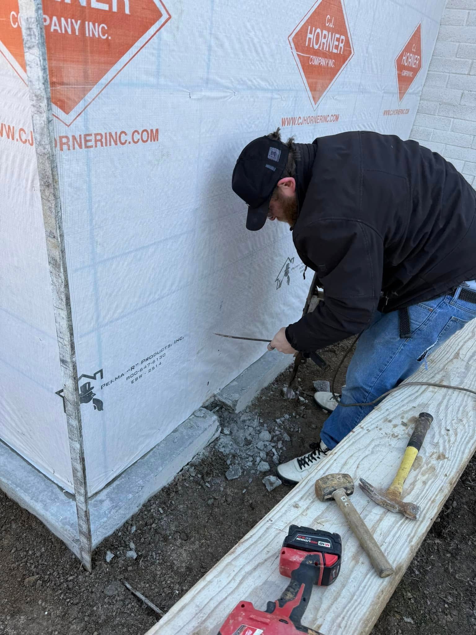 Worker sealing the base of a building wall with tools on the ground nearby.