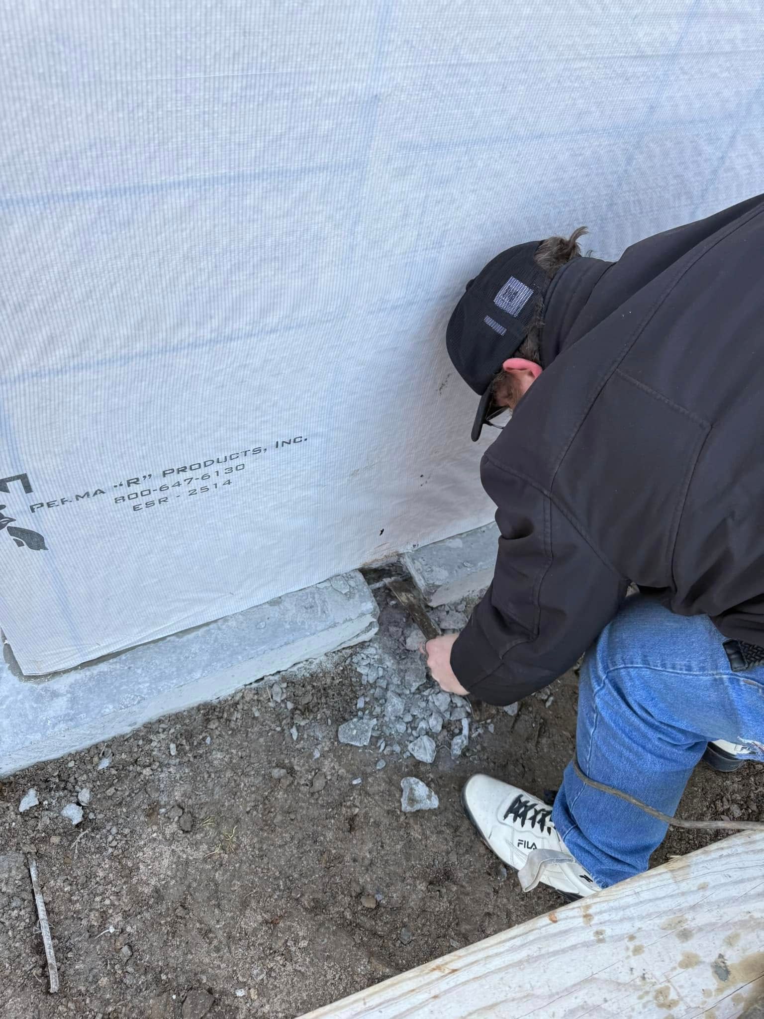 Person kneeling beside a house foundation, working near a white wrapped wall on bare ground.