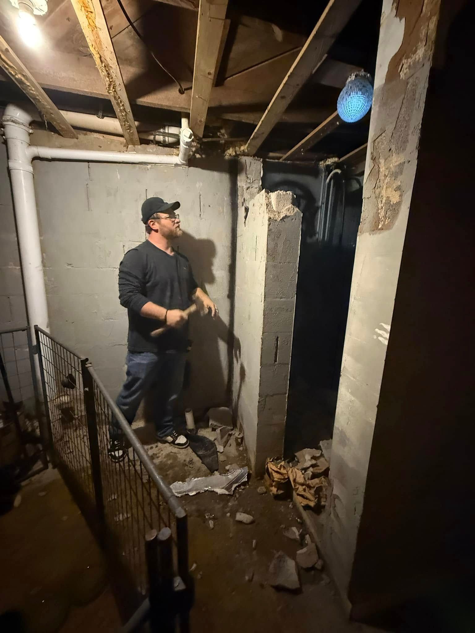 Person in a dark basement under exposed beams, looking up while holding a tool in a damaged stairwell.