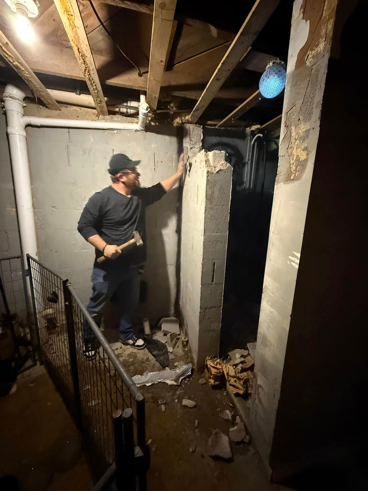 Person inspecting damaged wall and debris inside a dim, unfinished room with exposed ceiling beams