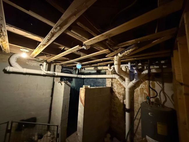 Basement utility room with exposed pipes, wooden joists, and a water heater in the corner.