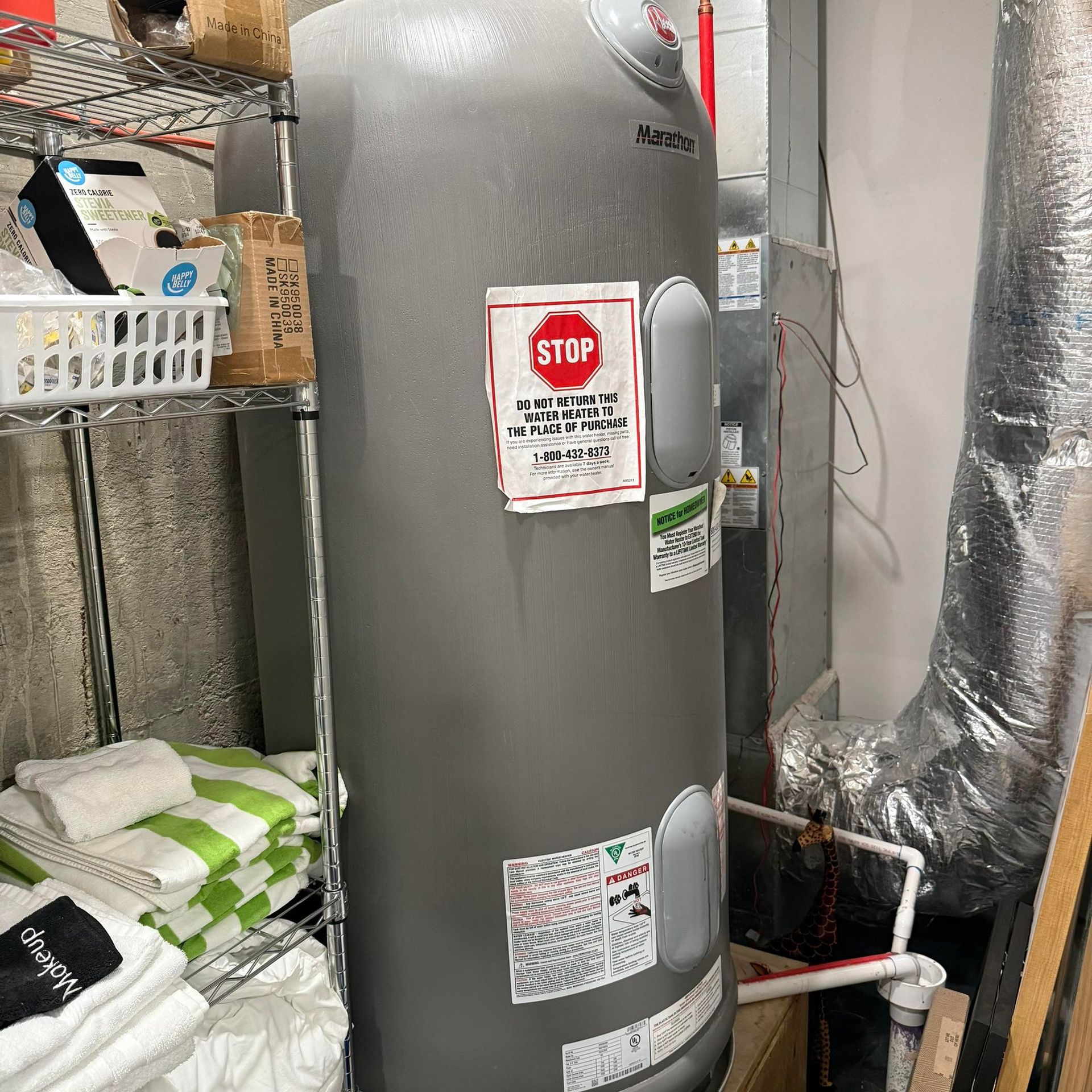 Gray water heater with warning labels in a utility/storage closet, next to shelves and insulated pipes.