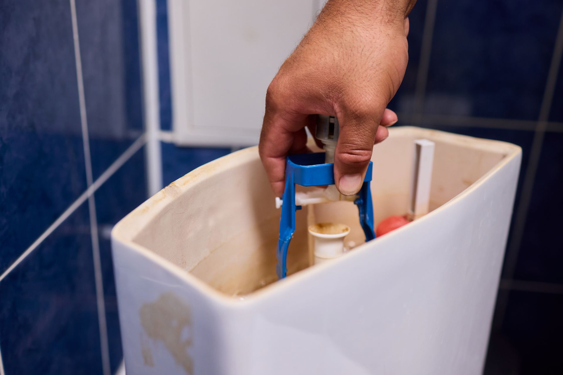 Hand filling a toilet tank with a blue bottle in a blue-tiled bathroom