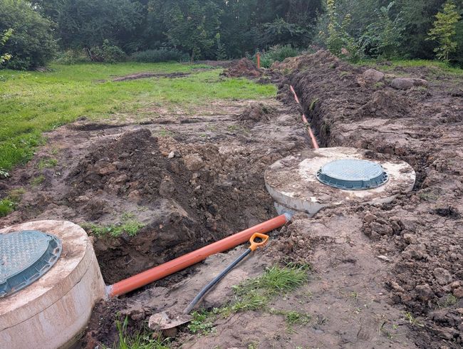 Excavated trench with two round concrete covers connected by orange pipe in a grassy outdoor area