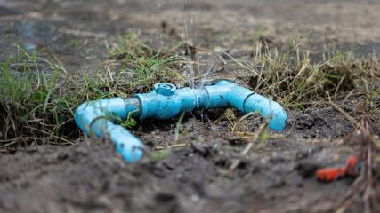 Broken blue pipe leaking on muddy ground with sparse grass and a small orange piece nearby