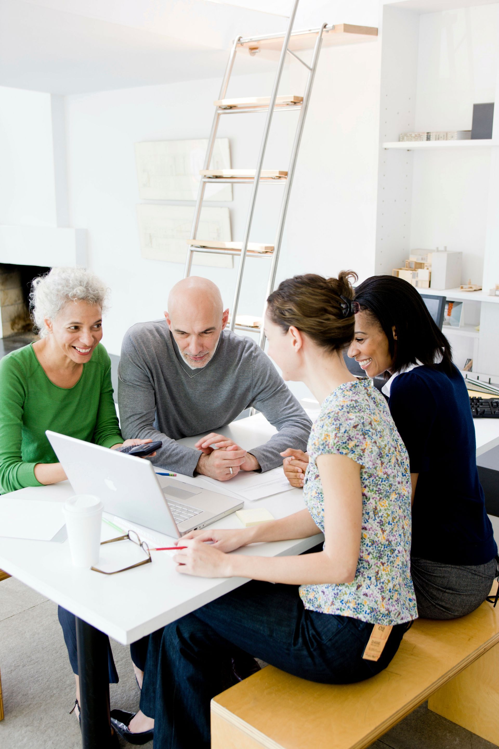 Four people seated at a white table, looking at a laptop. A woman points at the screen. Bright, airy room.