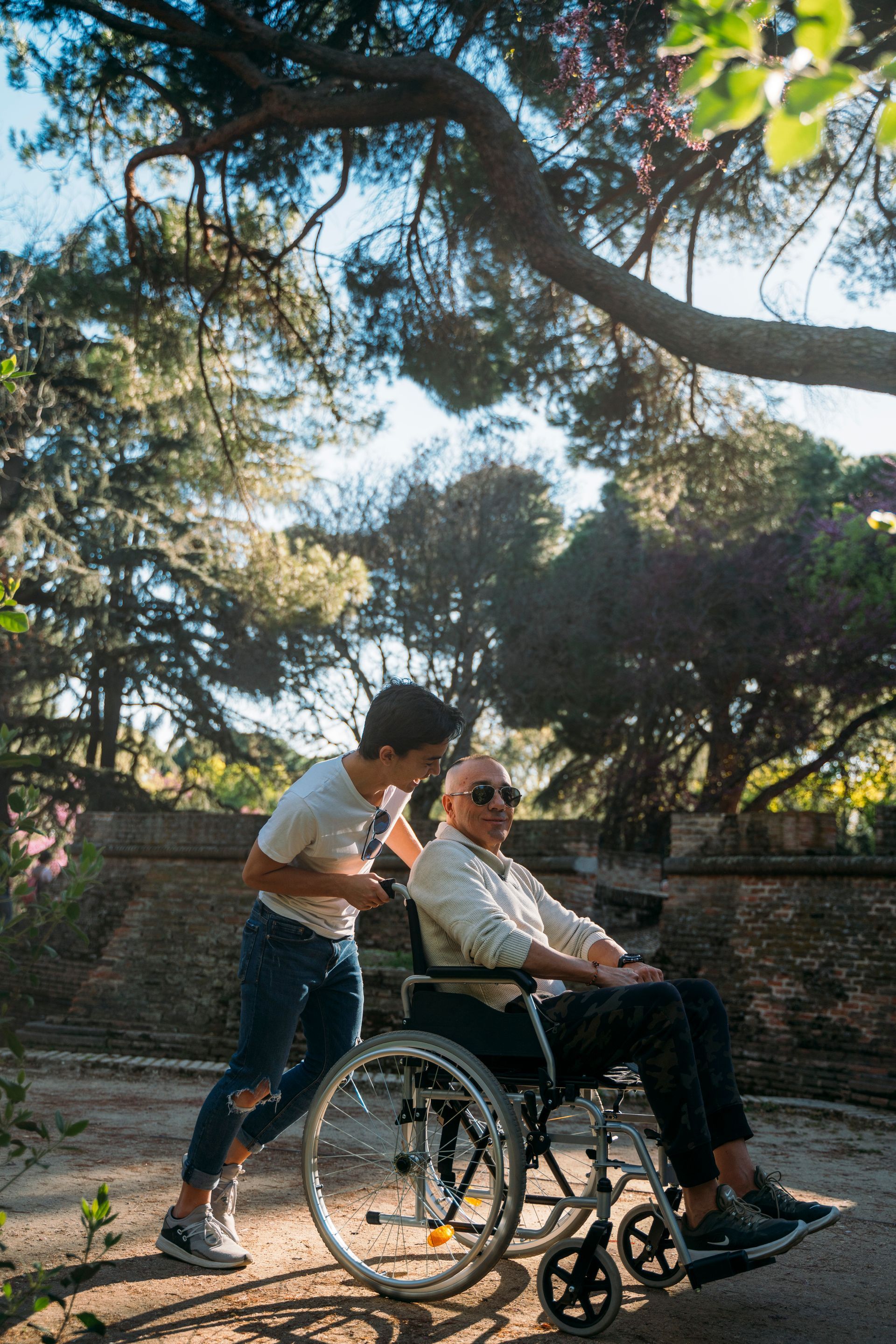 Young person pushing a wheelchair with an older person outdoors under a tree, sunlight shining through.