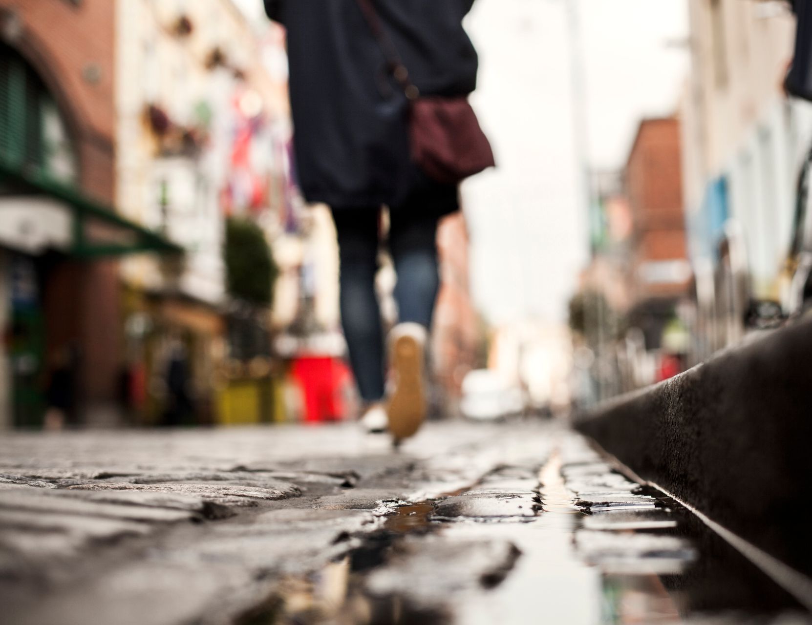 Person walking on a cobblestone street, brown shoes, maroon bag, blurred buildings.
