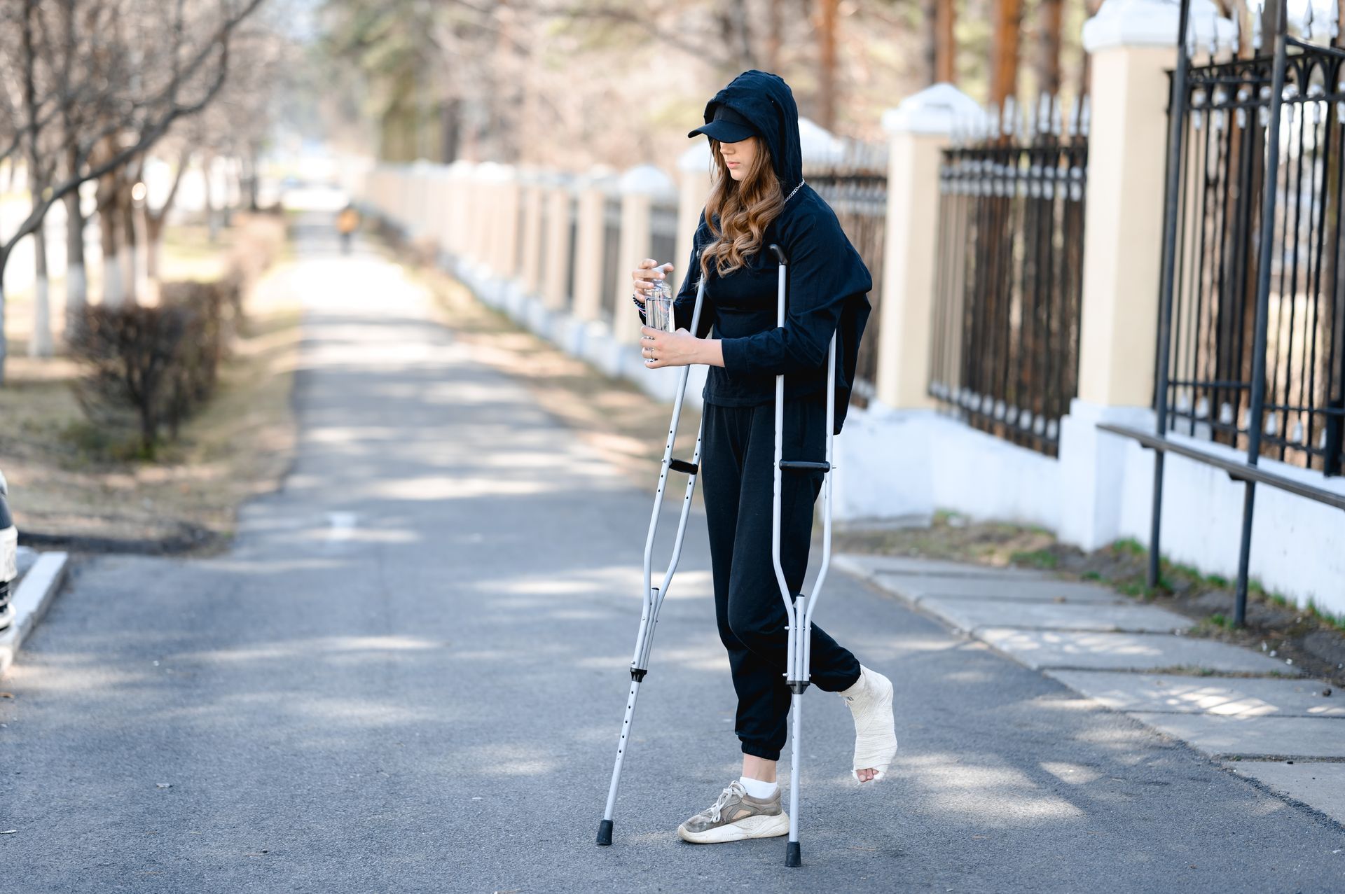 Woman with crutches walking on a path, foot in cast. Wearing black hoodie and pants. Outdoors, near a fence.