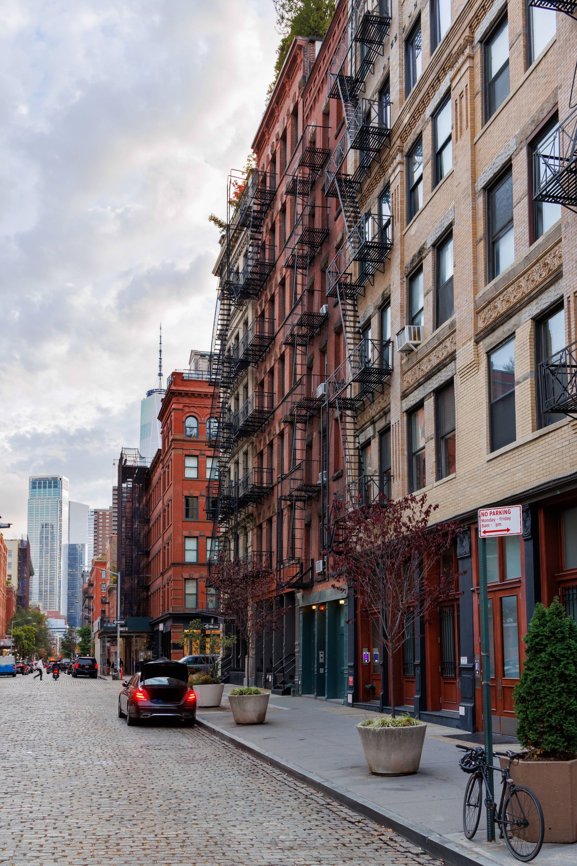 Cobblestone street in New York City with tall brick buildings, fire escapes, trees, and parked cars.