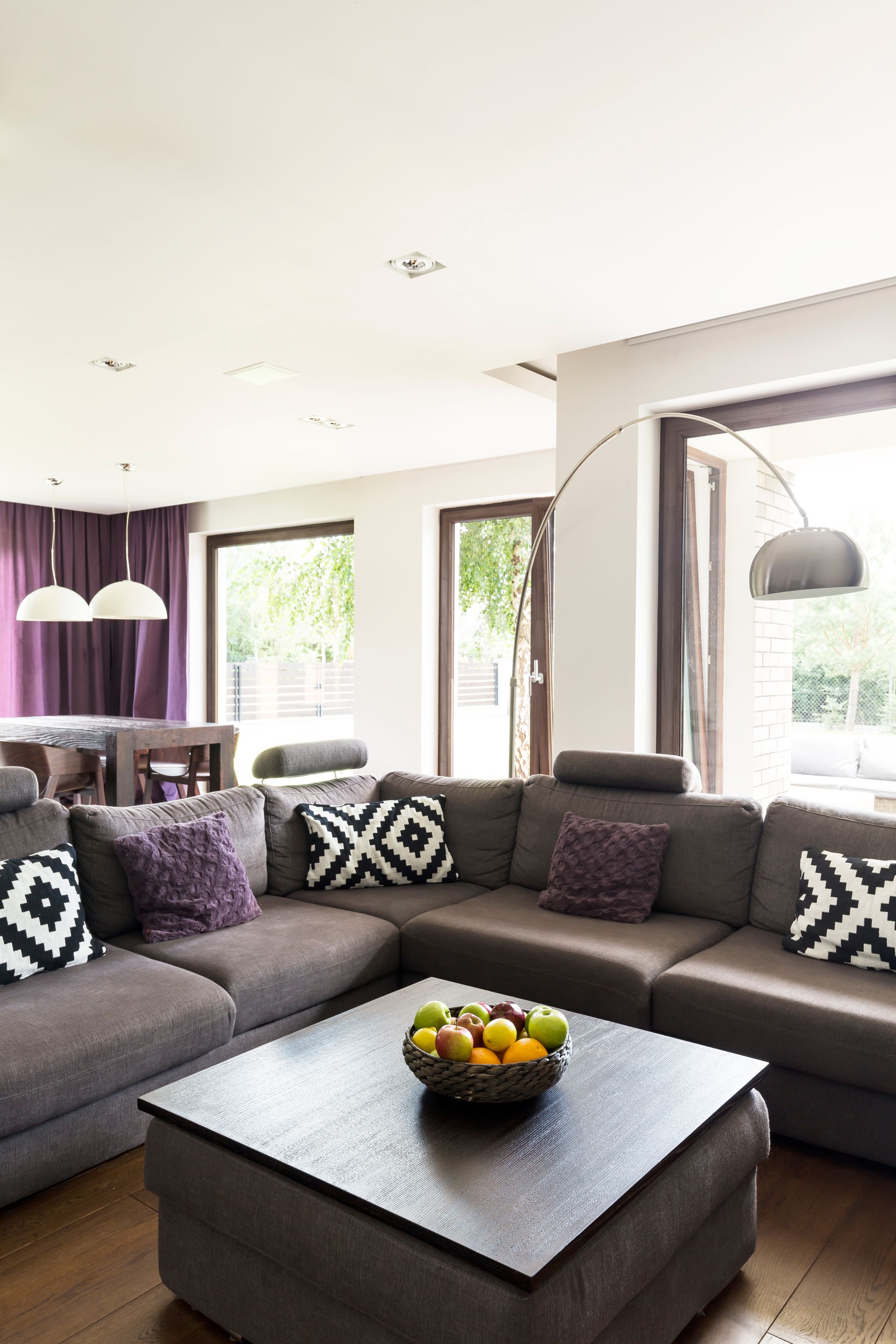 L-shaped gray sofa with patterned pillows, square coffee table with fruit bowl, near window with arched lamp.