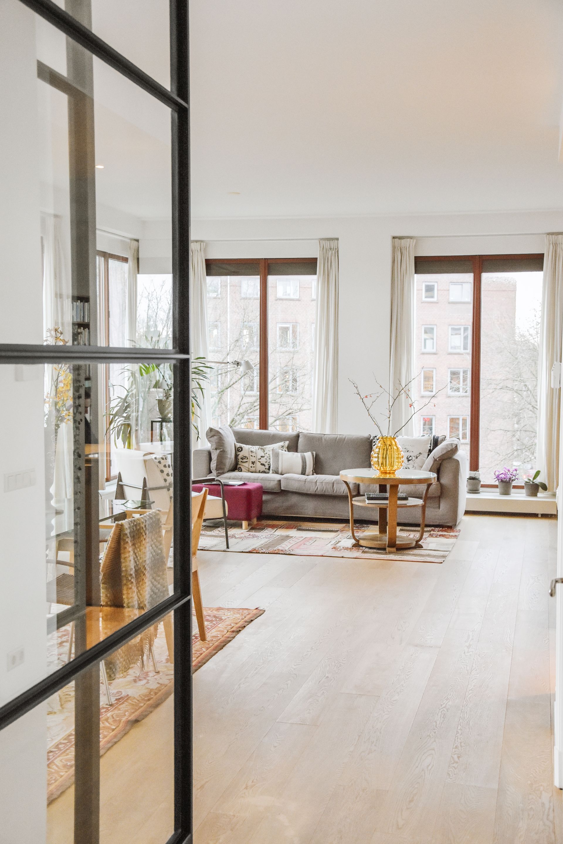 Living room with a gray couch, wooden floors, and large windows. View through a glass door.