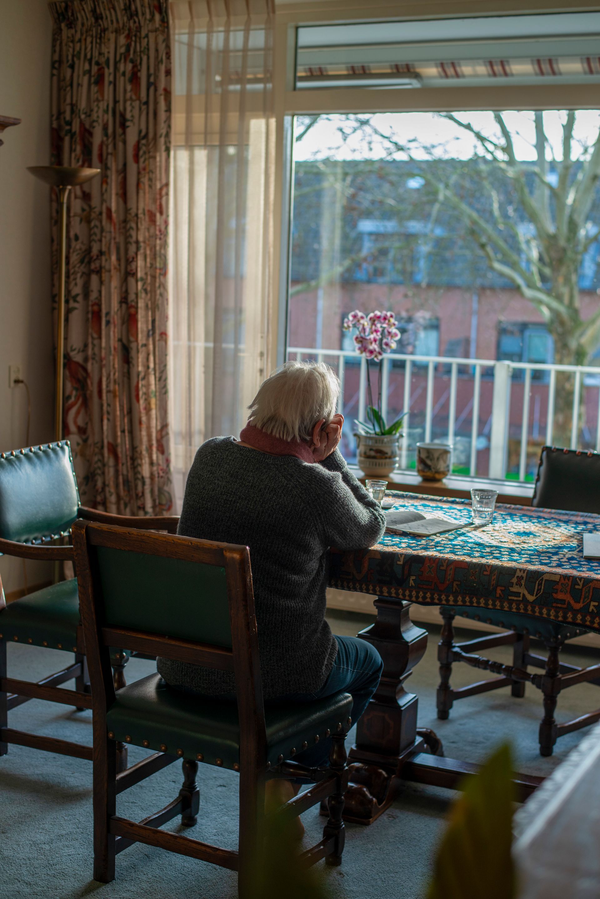 Person sits at table, looking out a window. Floral curtains, table with rug, plants, view of building.