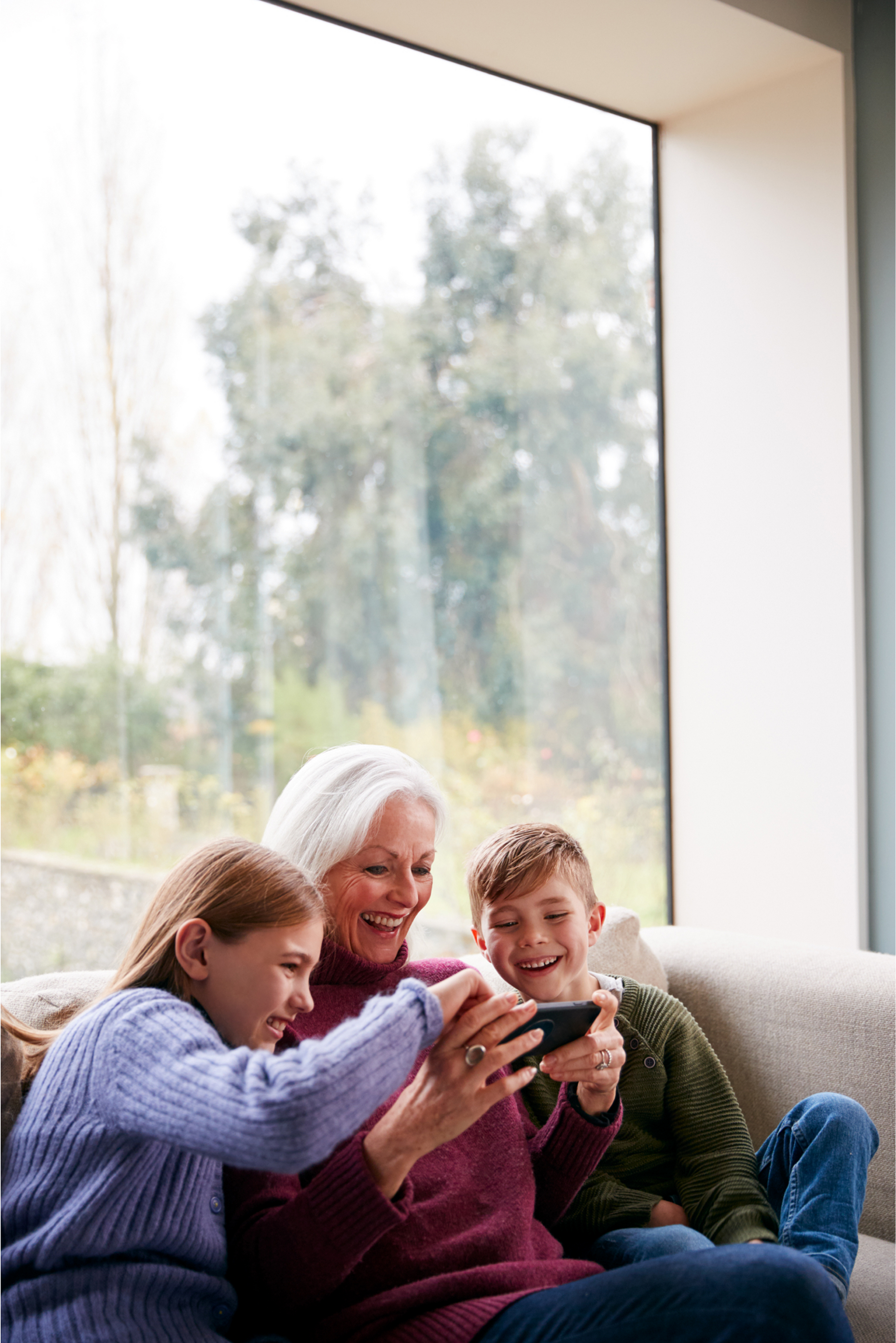 Woman and two children on couch looking at a phone screen. Large window and trees in the background.