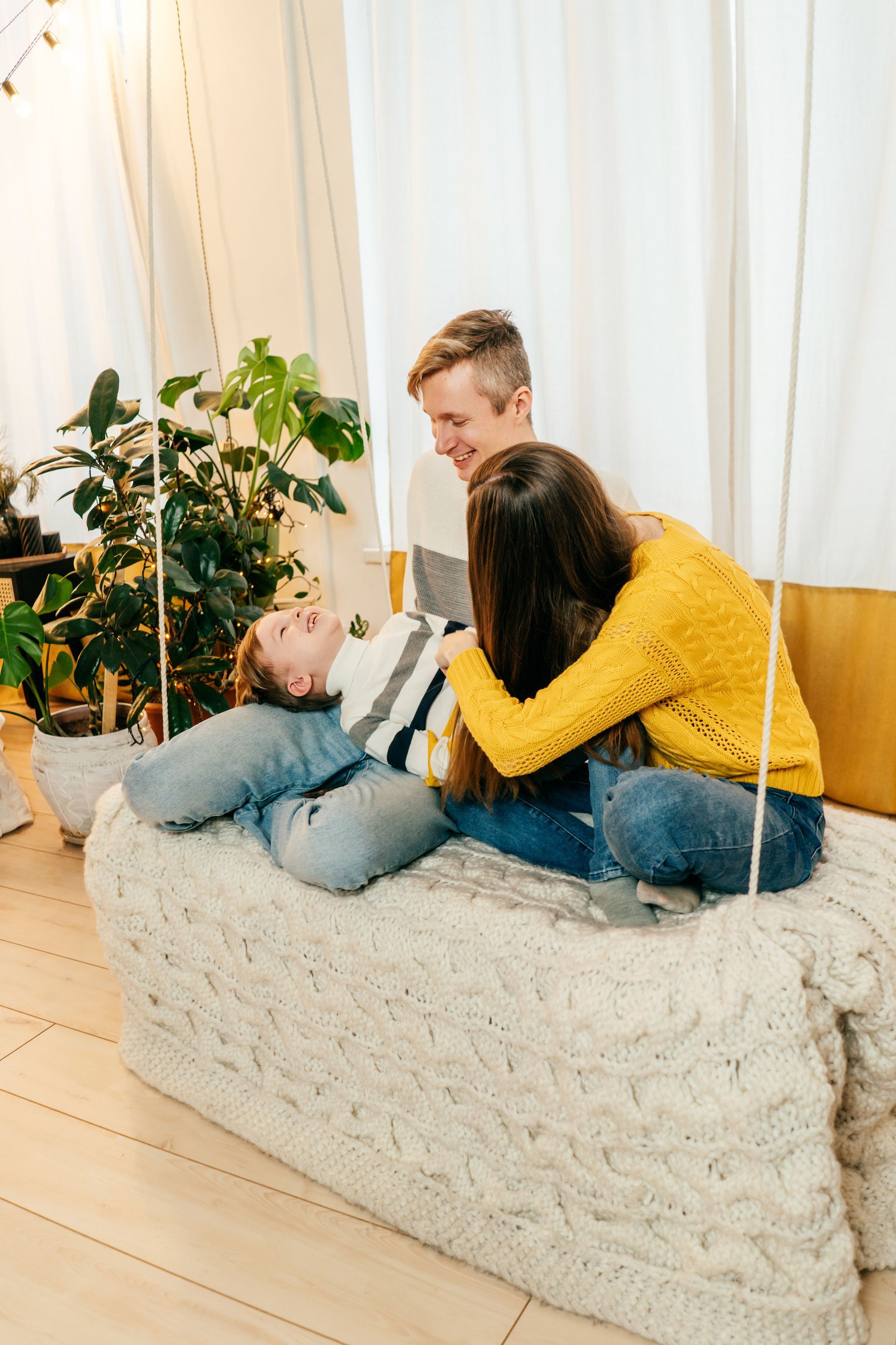 Family on swing seat, laughing. Child on parent’s lap. Cozy interior with plants, draped curtain.