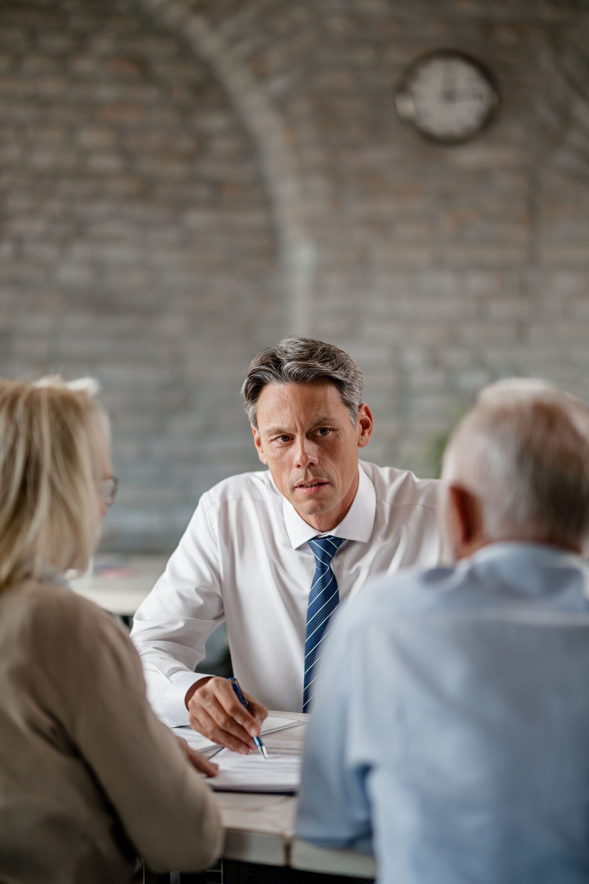 Man in tie reviews paperwork with an older couple seated at a table. Brick wall and clock visible.