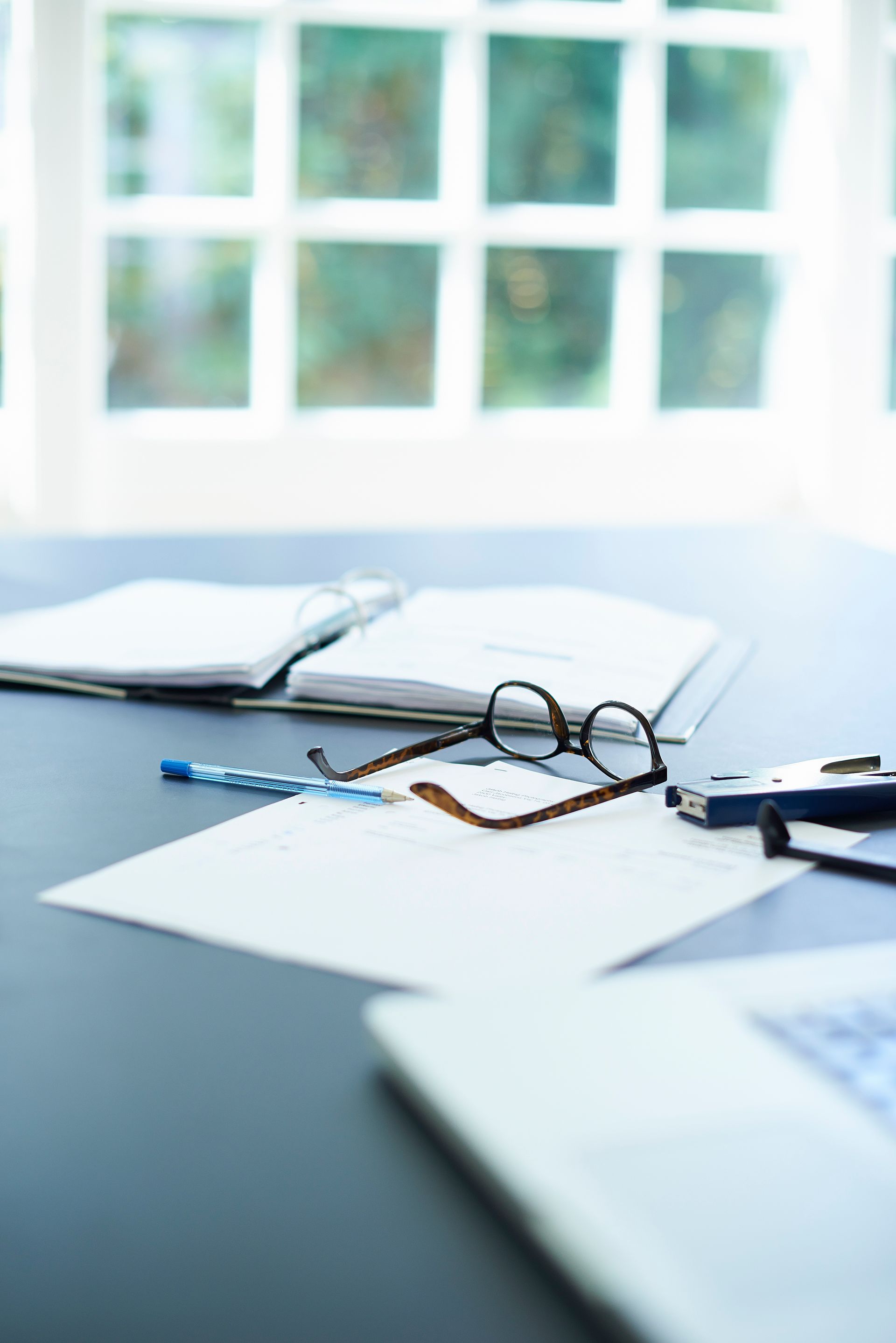 Desk with papers, glasses, pen, and laptop in front of a window.
