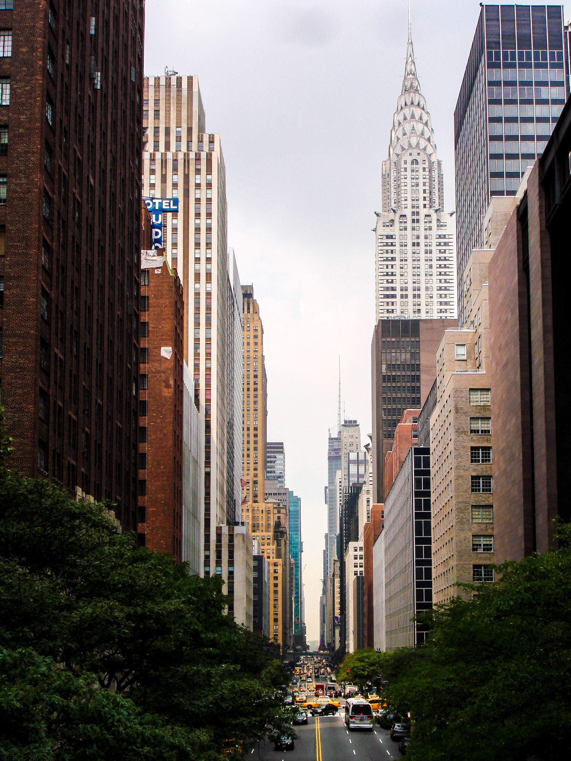 New York City street view with the Chrysler Building in the background. Buildings line both sides, and cars drive on the street.