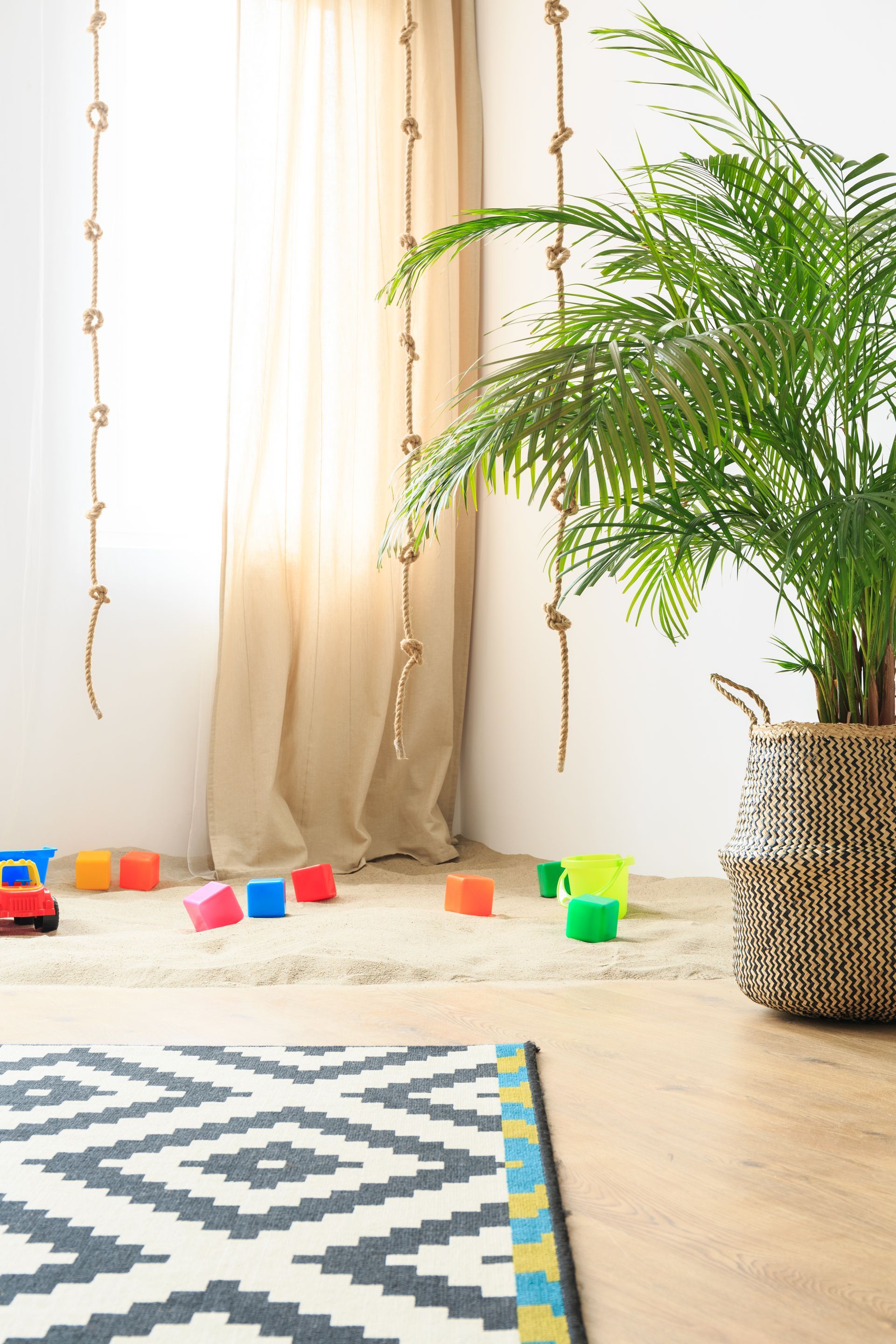 Room with a potted palm, colorful toys on the floor, rope decorations, and a geometric rug.