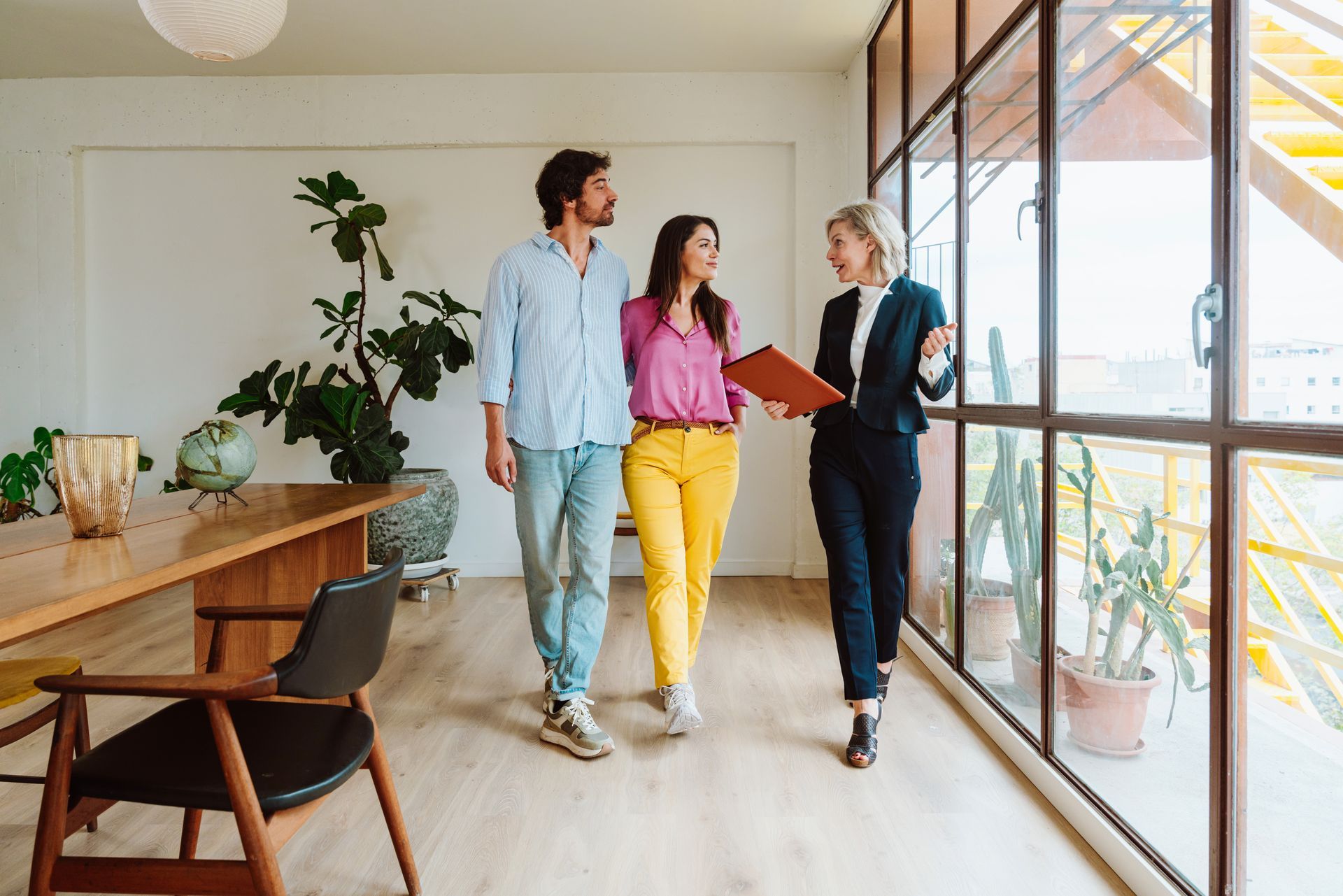 A real estate agent showing a couple a brightly lit apartment with a long wooden table and large windows.
