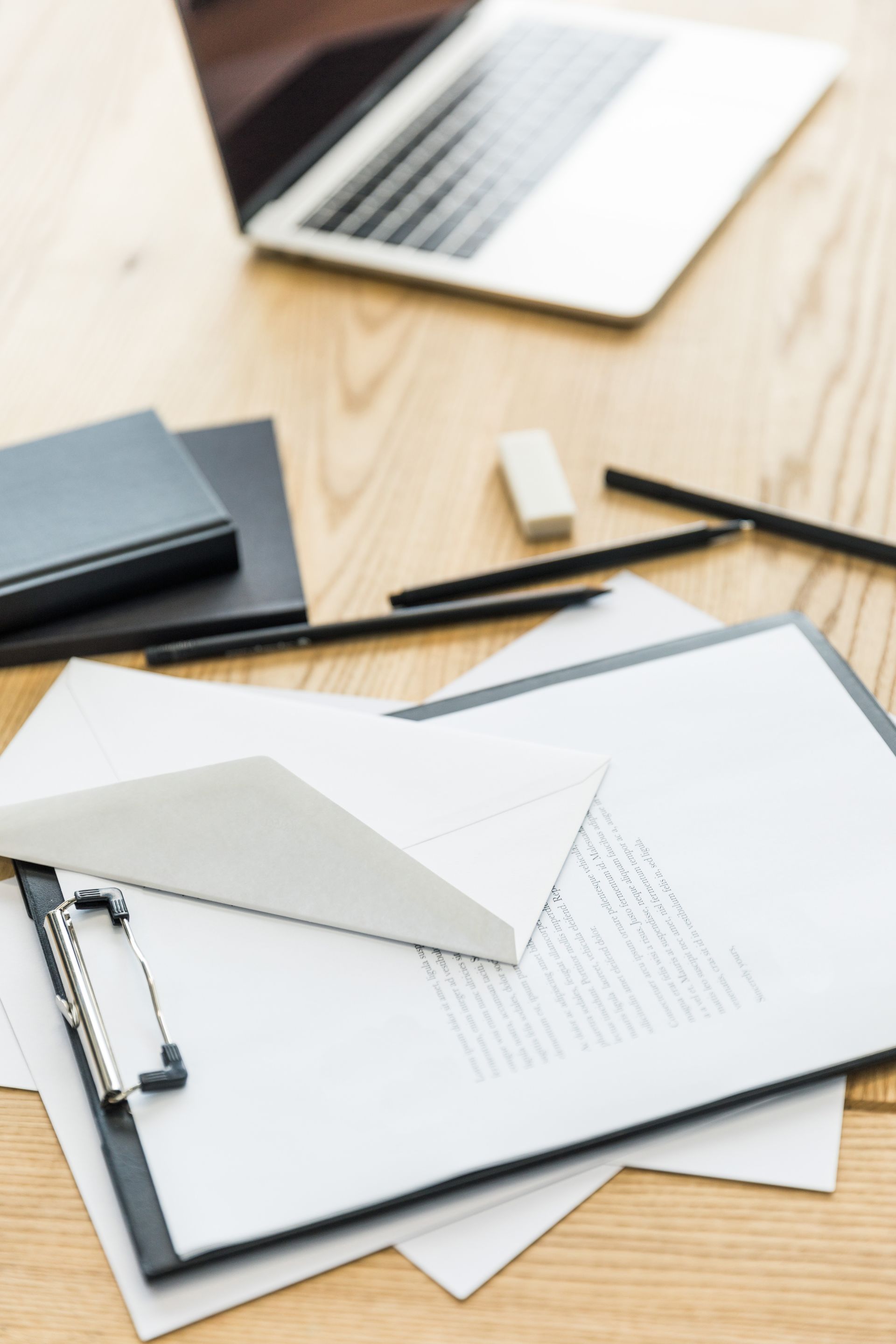Laptop, clipboard with papers, stationery, and notebooks on a wooden desk.