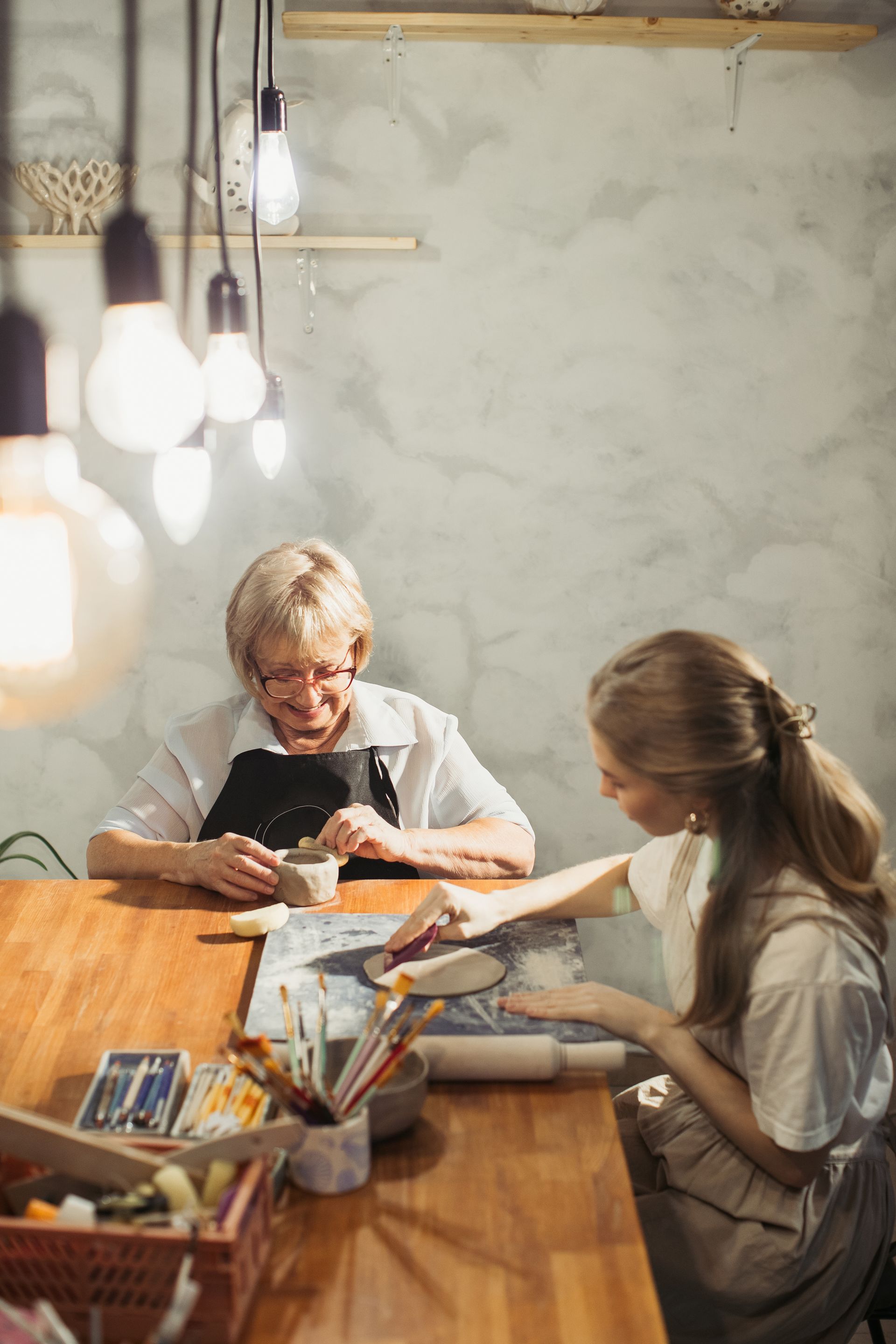 Woman and person working on pottery at a table in a workshop, under hanging lights.