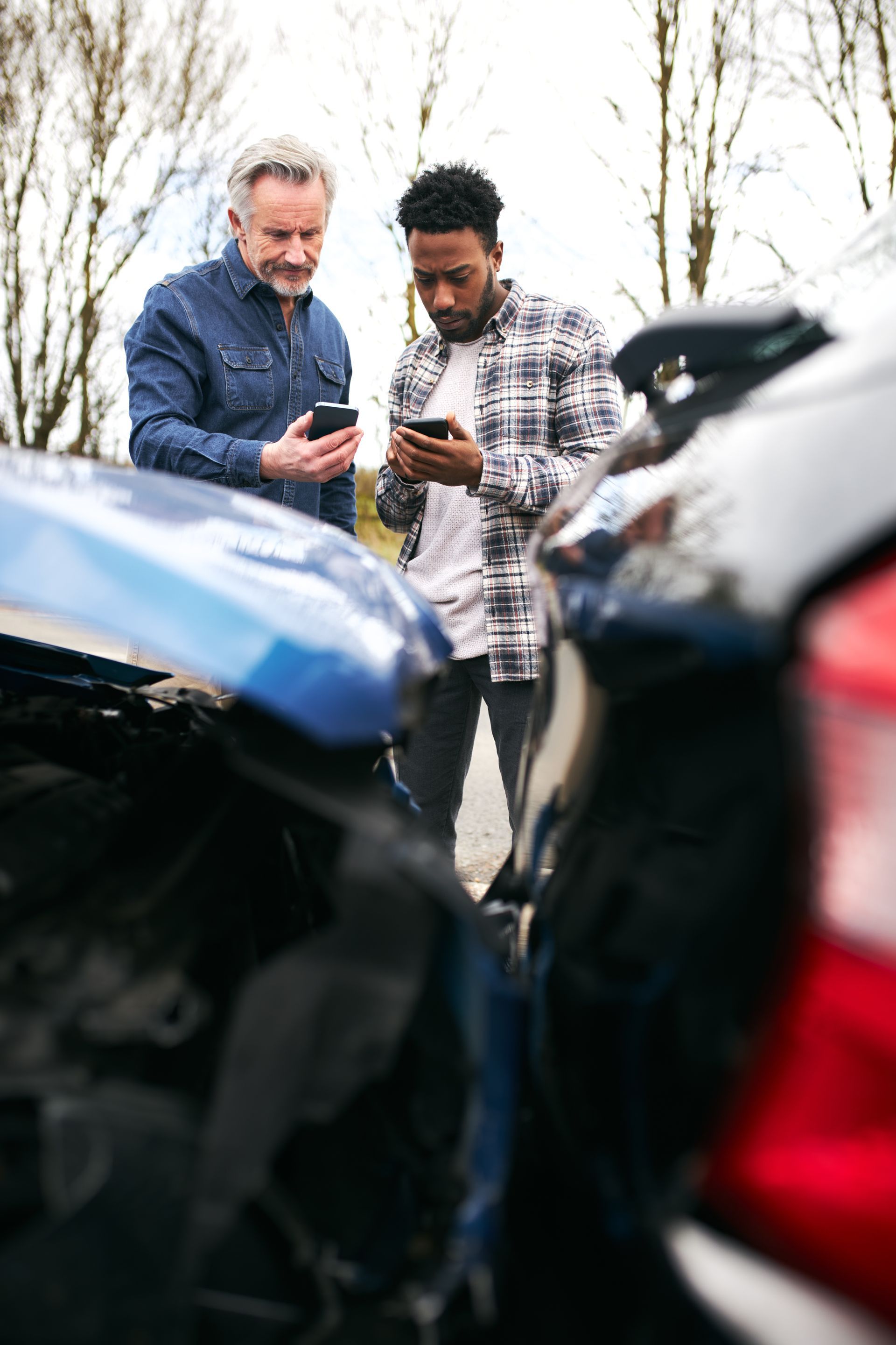 Two men looking at their phones near damaged cars after a collision.