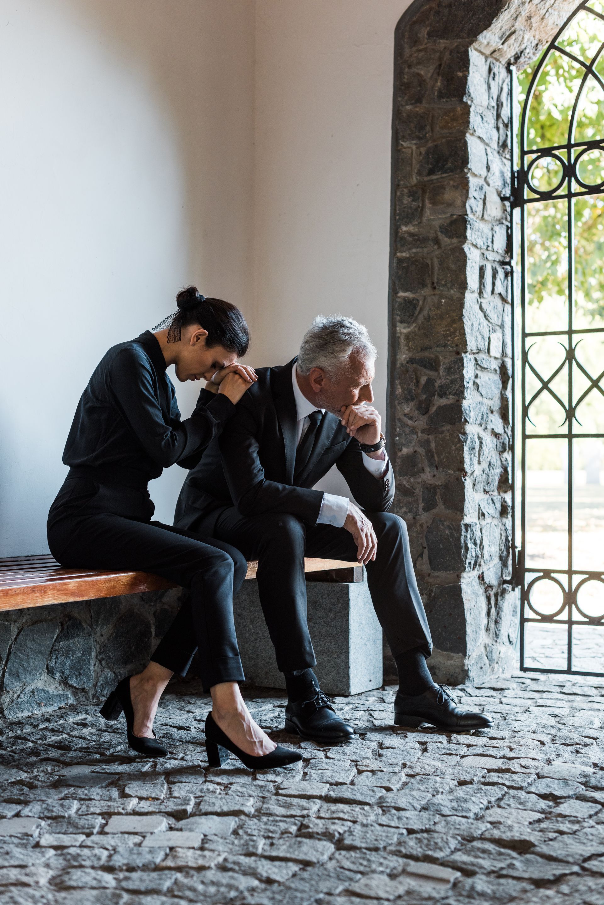 Woman comforts a man on a stone bench near a building entrance, both in black clothing, somber expressions.