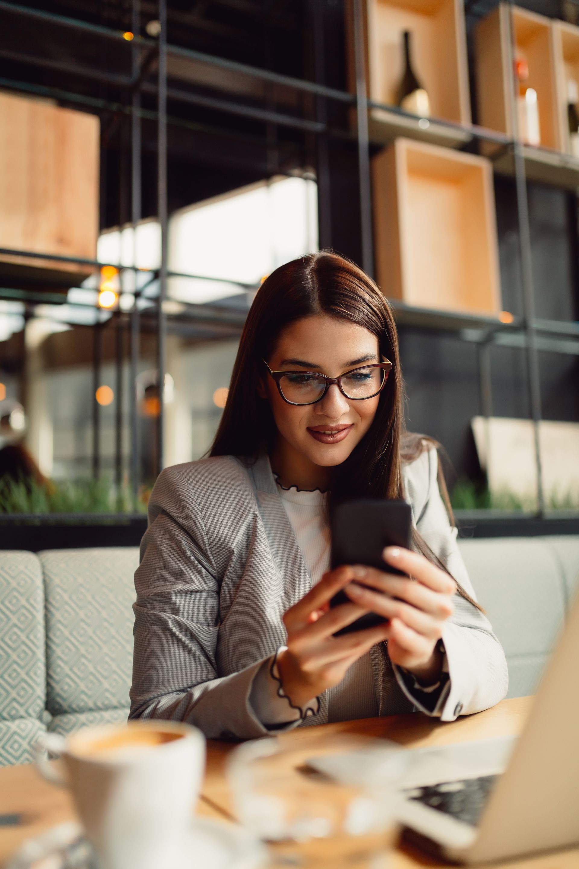 Woman wearing glasses looking at phone, sitting in cafe with laptop and coffee.