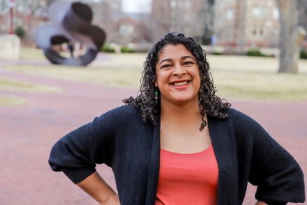 Smiling woman in a coral top and black cardigan standing outdoors on a campus walkway