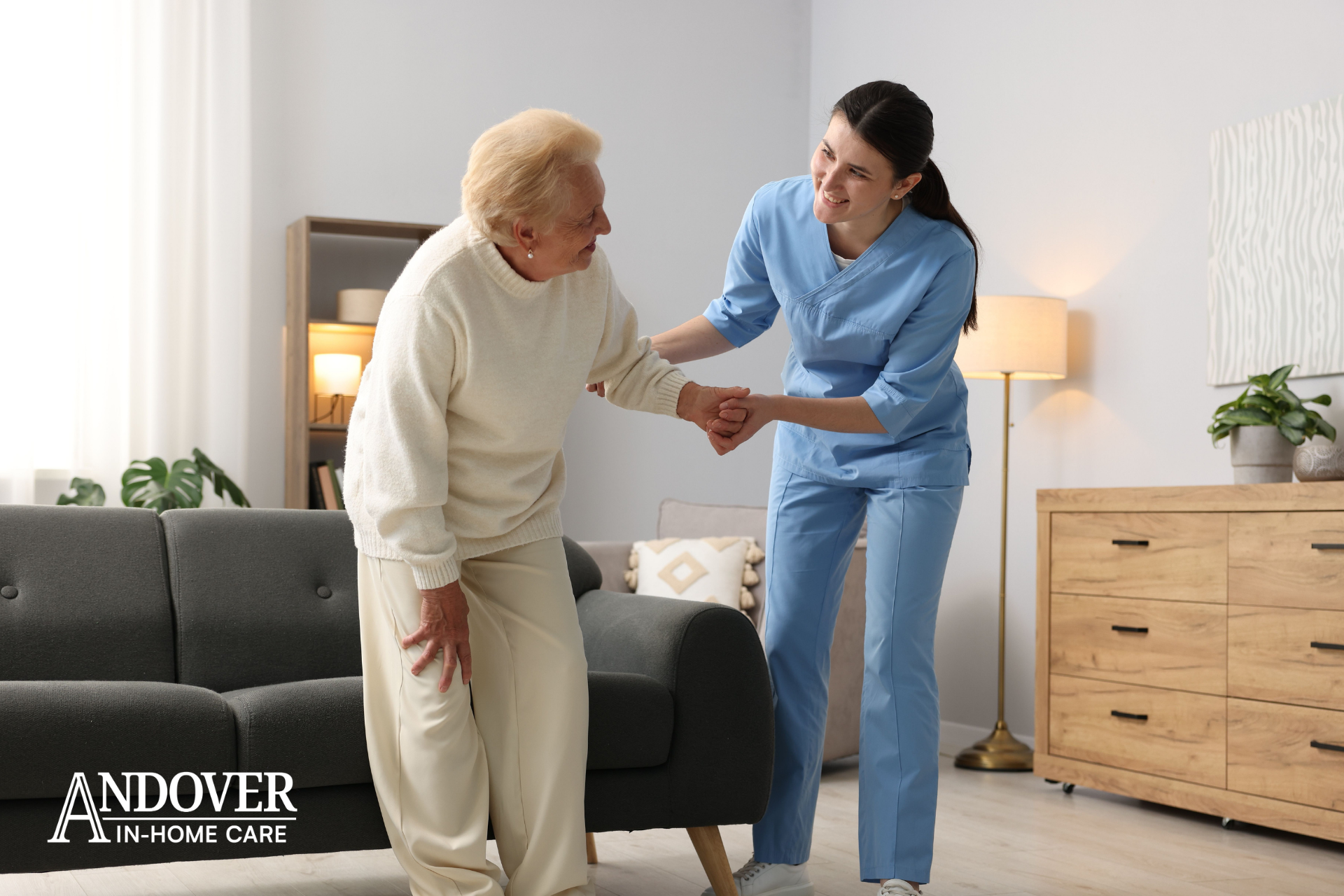 Caregiver helping an elderly person stand in a living room with a sofa and dresser.