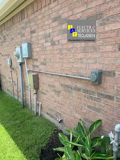 Exterior brick wall with electrical meters, conduit, and a small utility sign above a grassy strip.