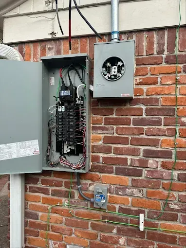 Open electrical panel and meter on a brick exterior wall with exposed wiring and conduit.