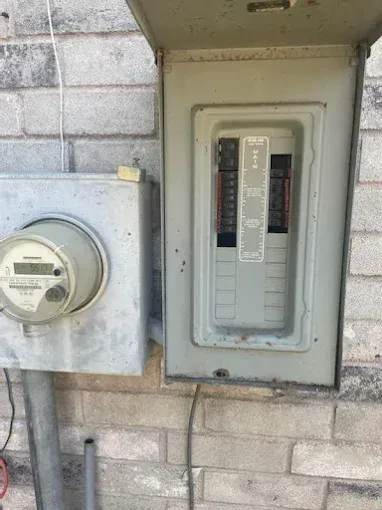 Electric breaker panel beside a utility meter on a cinder block wall.