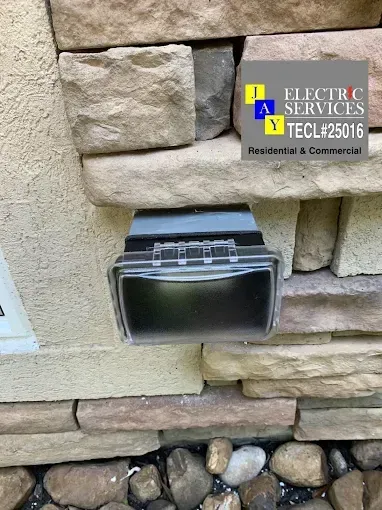 Outdoor wall electrical outlet under stone veneer, with an uncovered black box and nearby utility label.