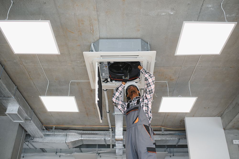 Worker in a gray shirt repairing an open ceiling electrical panel in a concrete room with bright square lights