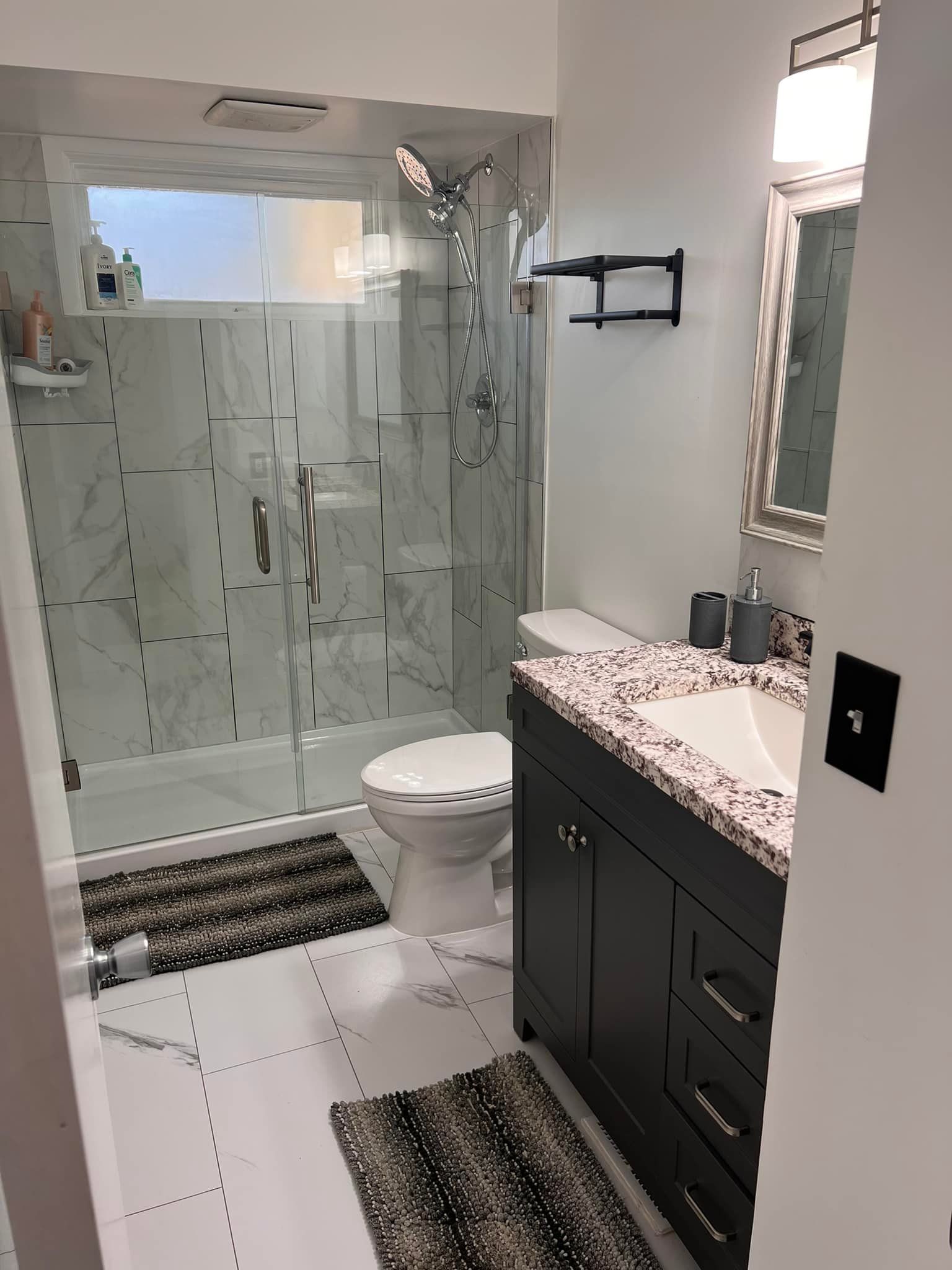 Small bathroom with glass shower, toilet, vanity, and striped rugs on white tile flooring.