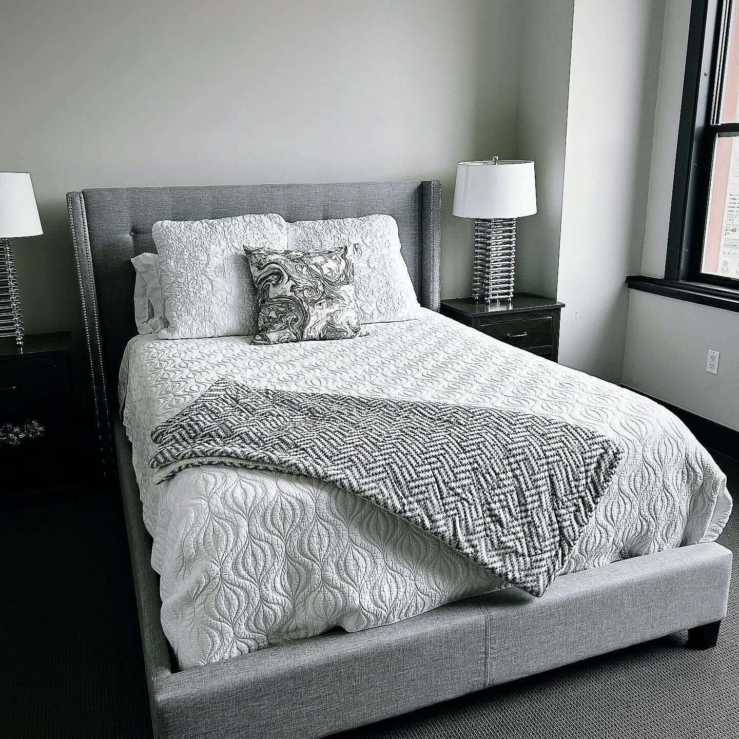 Gray upholstered bed with white bedding and bedside lamps in a bright bedroom