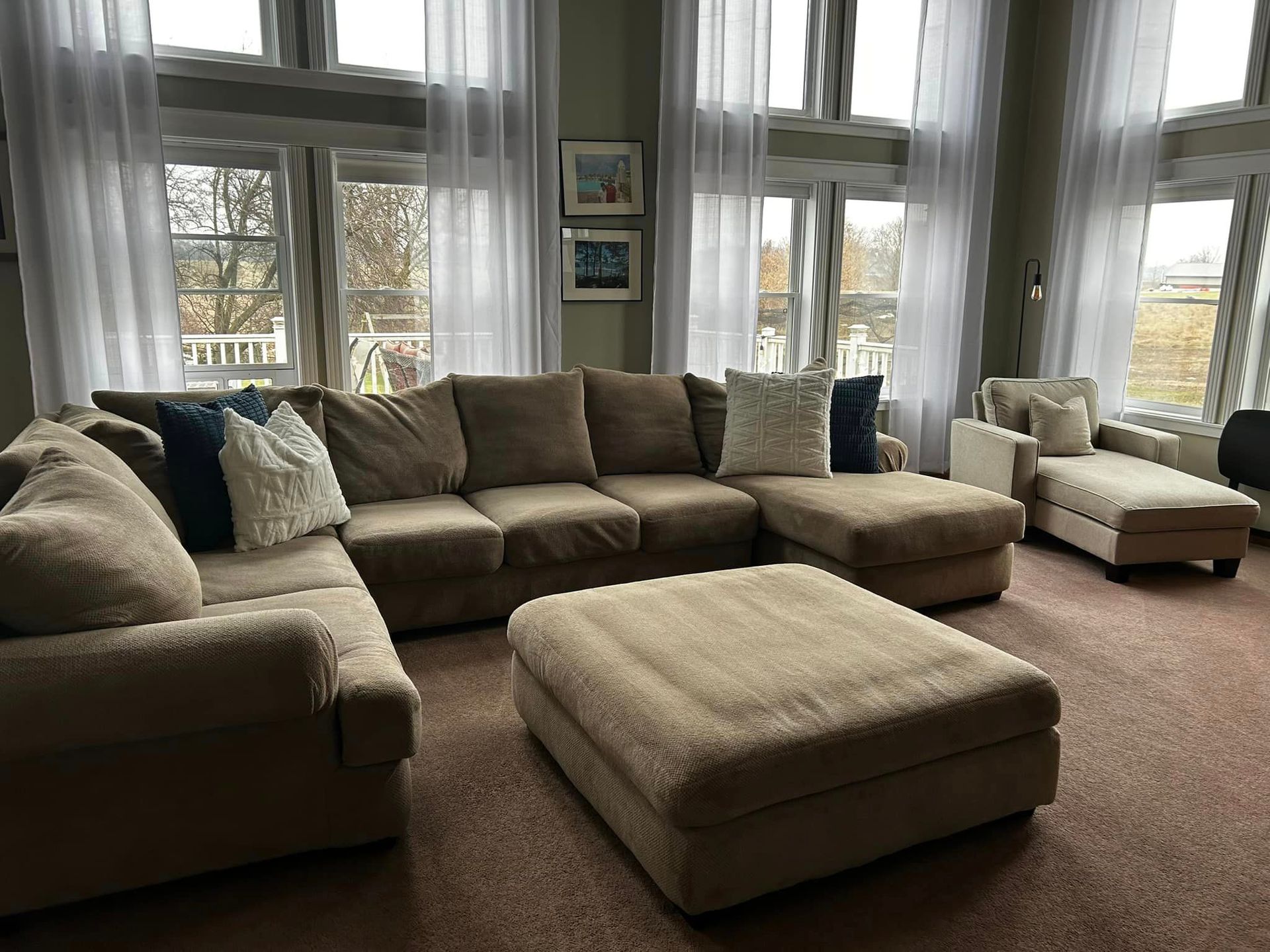 Bright living room with beige sectional sofas, ottoman, and large windows overlooking trees.