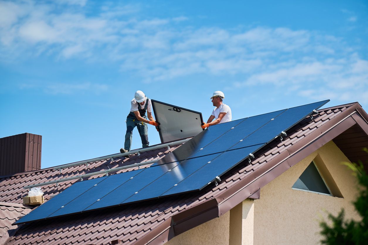 Two workers installing solar panels on a brown roof under a blue sky.