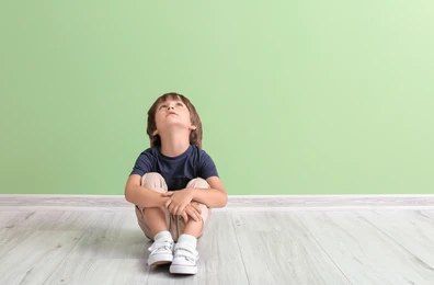 Child sitting on a light wood floor against a pale green wall, looking upward with knees hugged close — PoDDSS in Cairns, QLD