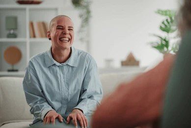 Woman with shaved head laughs while sitting on a couch. Indoor setting with a plant and bookshelf visible.— PoDDSS in Hyde Park, QLD