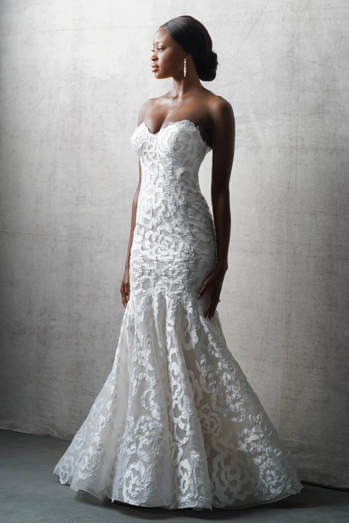 A smiling person posing in a floor-length, white, sleeveless fitted gown against a light wall and stone path.
